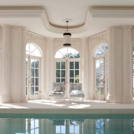 Janine Stone & Co residential architecture — classical indoor poolhouse with stone arched French windows, Doric columns, white-glass pendant and pair of poolside daybeds.